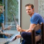 A man in a blue t-shirt cleaning large glass pane with a squeegee indoors.