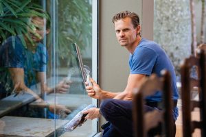 A man in a blue t-shirt cleaning large glass pane with a squeegee indoors.